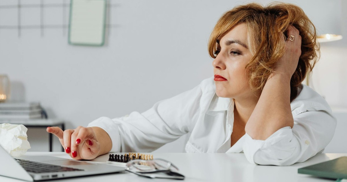 Frustrated woman at desk, using laptop with expression of stress. Indoors office environment.