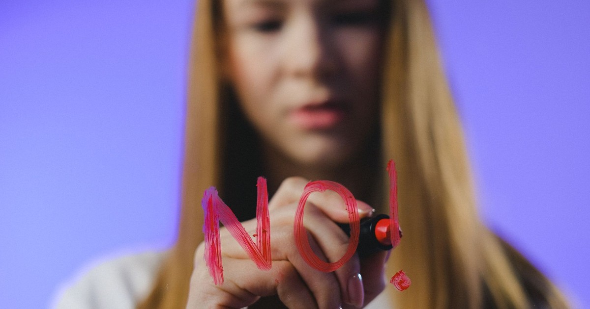 A teenage girl writes "No!" in bold red letters on a glass surface, against a blue background.
