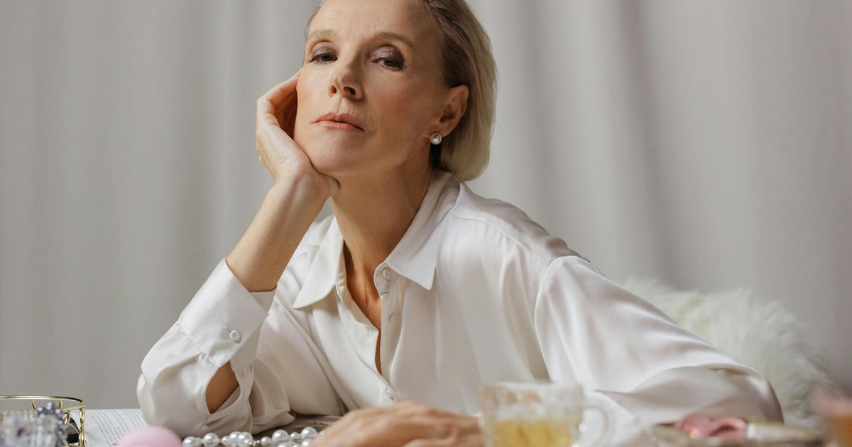 A poised senior woman in a white shirt enjoying tea, surrounded by elegant pearls.