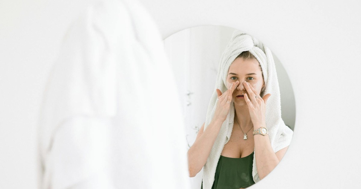 A woman in a towel performing her skincare routine in front of a bathroom mirror.