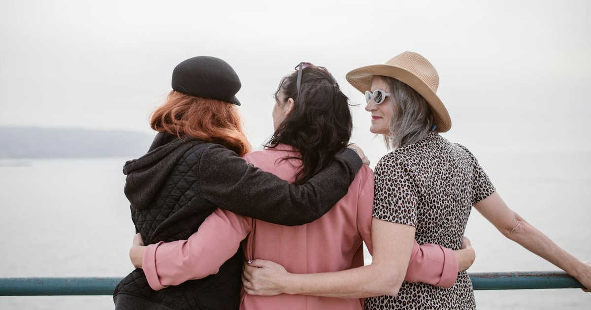 Three women, seen from behind, embracing by a seaside railing, symbolizing friendship.