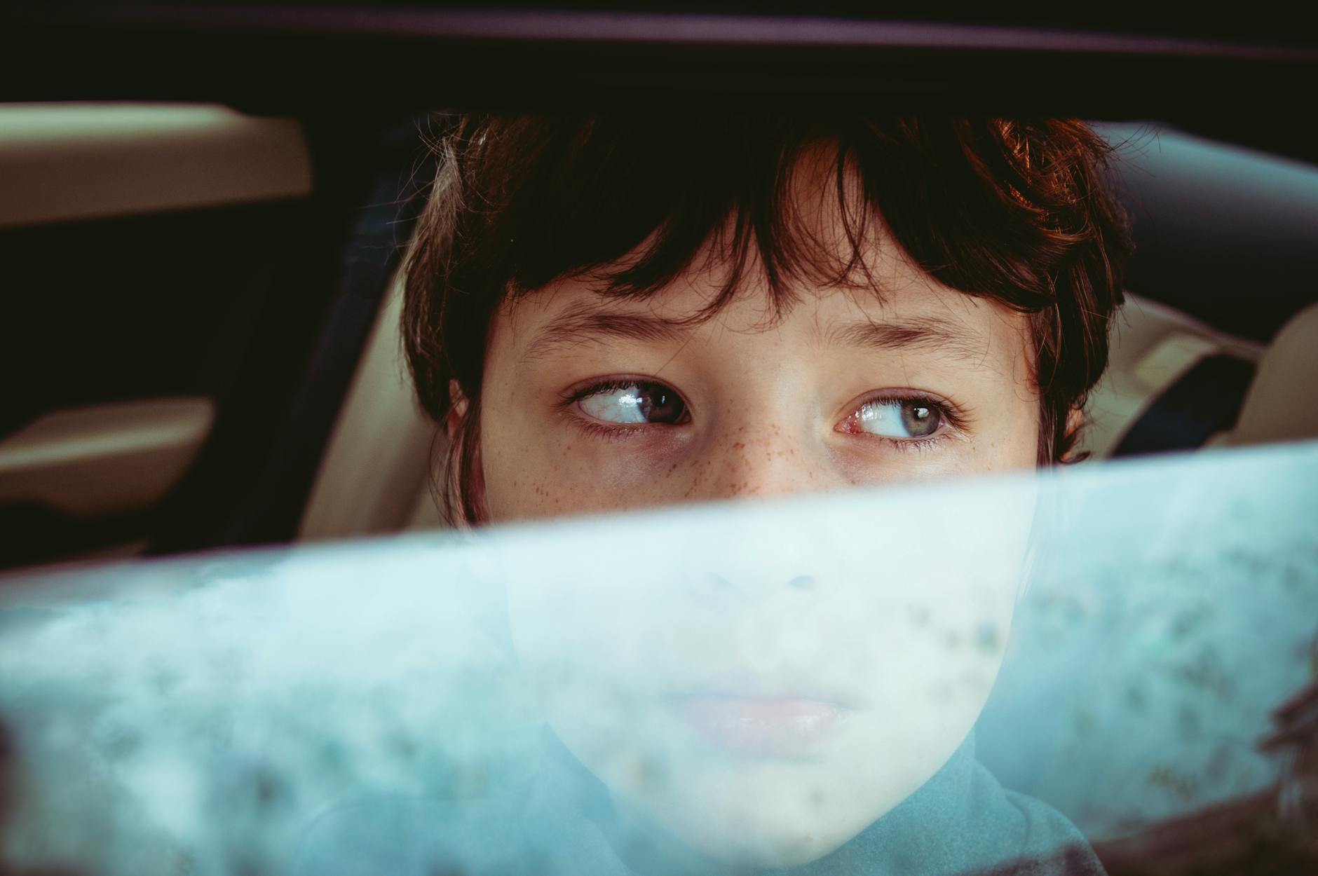 A young boy with blue eyes and brown hair peers thoughtfully through a car window.
