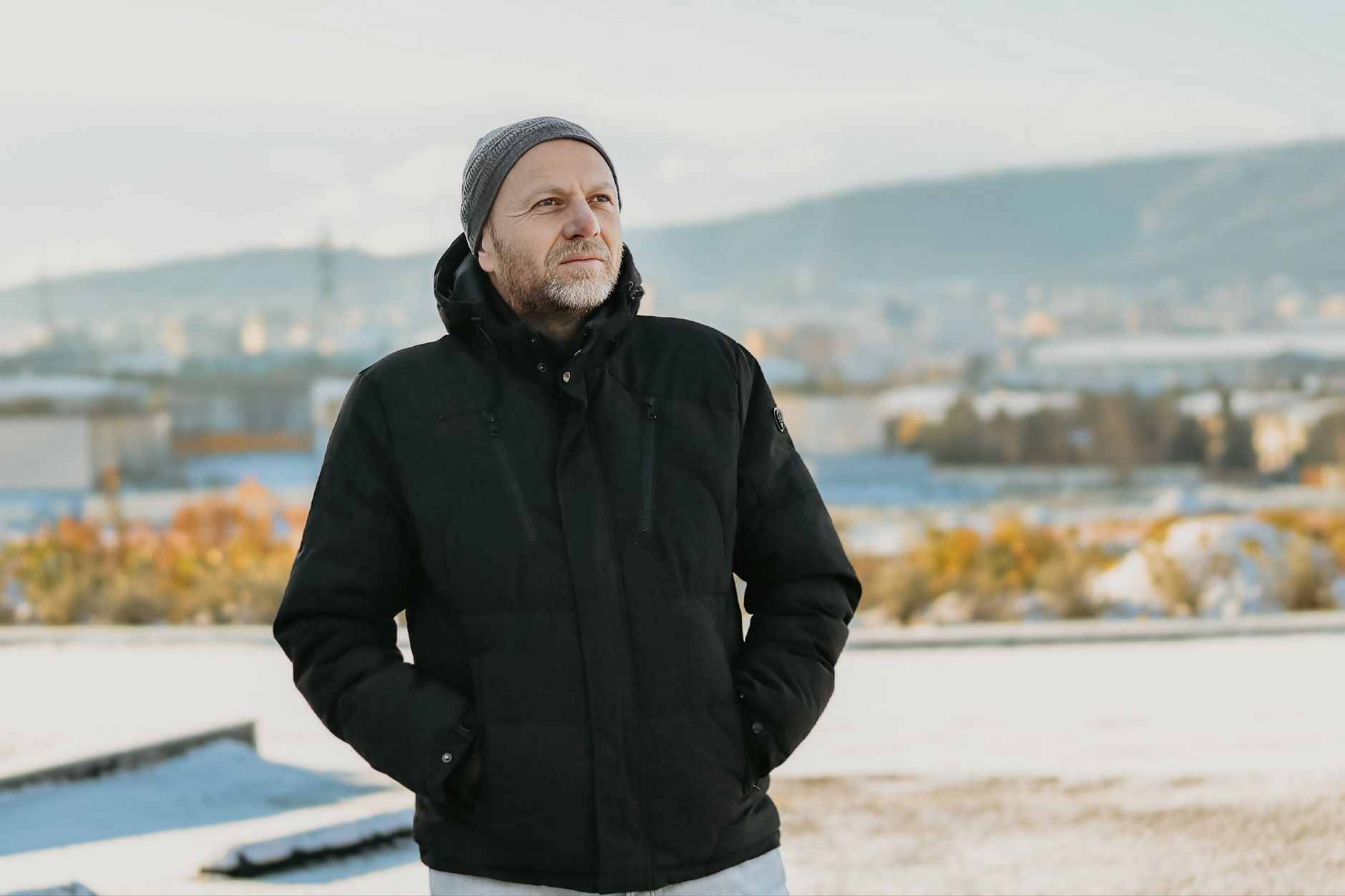 A middle-aged man in a black jacket and beanie outdoors in a winter setting, looking into the distance.