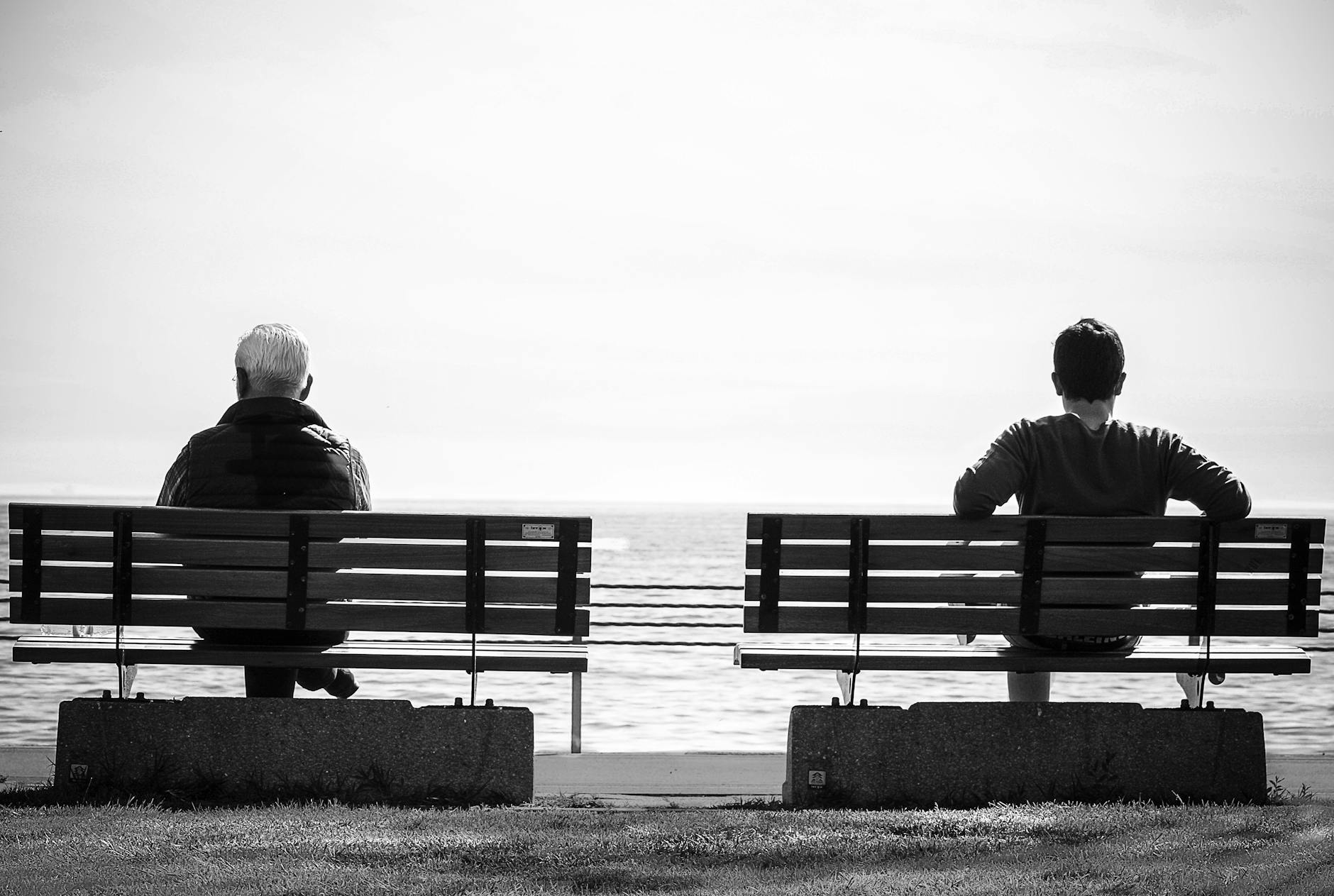 Two people sit on separate benches by the sea, captured in black and white.