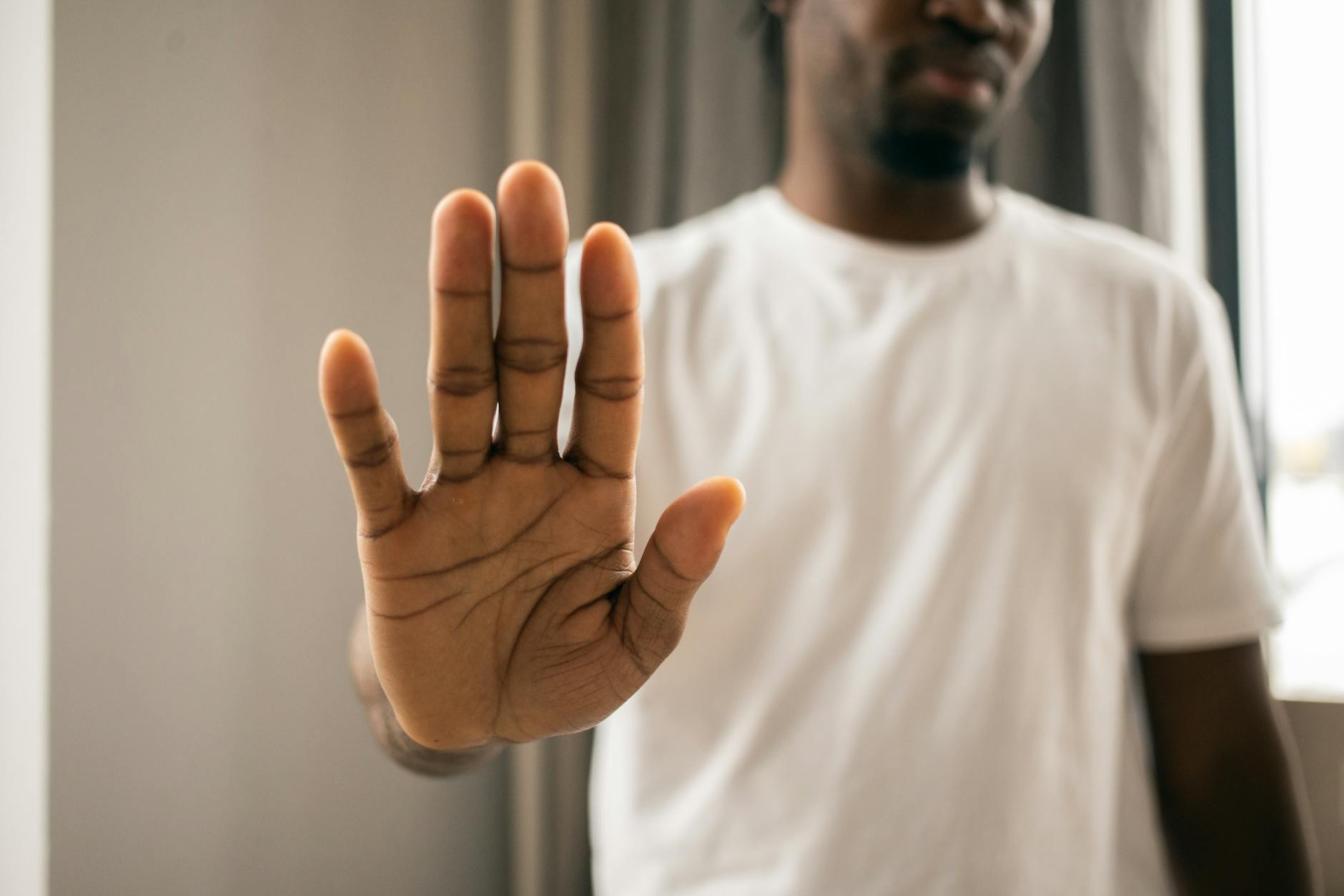 Crop anonymous African American male in casual white t shirt showing stop sign with hand while standing in light room