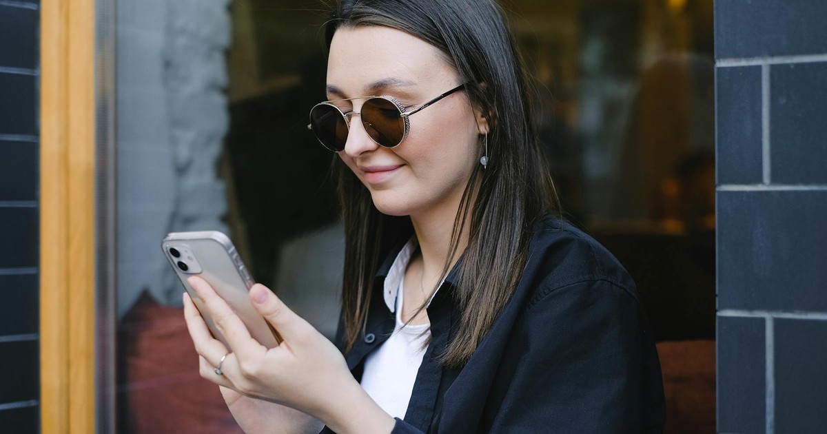 A young woman with sunglasses smiles while using her smartphone outdoors.