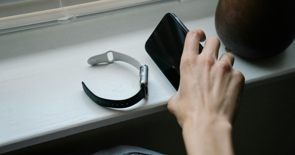 From above of crop anonymous person taking cellphone placed on windowsill near smart watch while waking up in morning