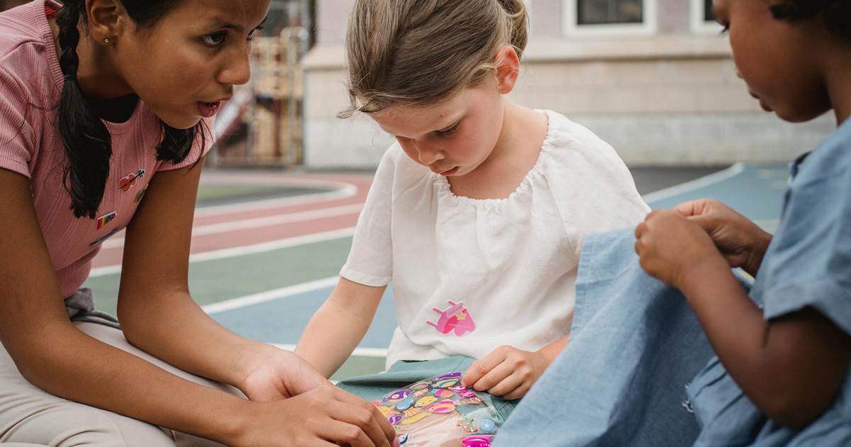 Three diverse children playing with stickers on an outdoor playground.