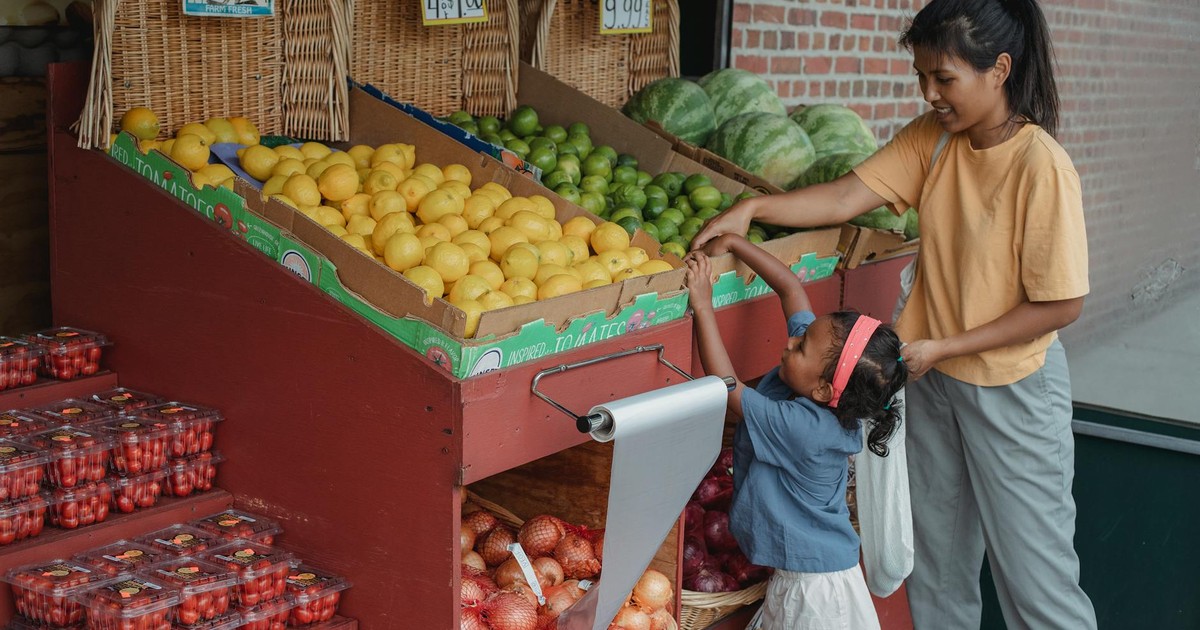 Positive Asian woman selecting ripe green apples with support of daughter while standing at street market with reusable bag