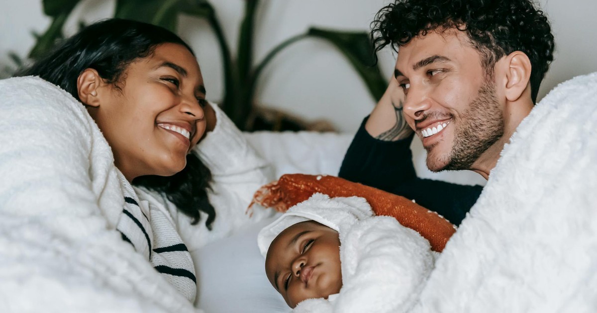 Positive ethnic couple in casual clothes lying near baby in comfortable bed and looking at each other