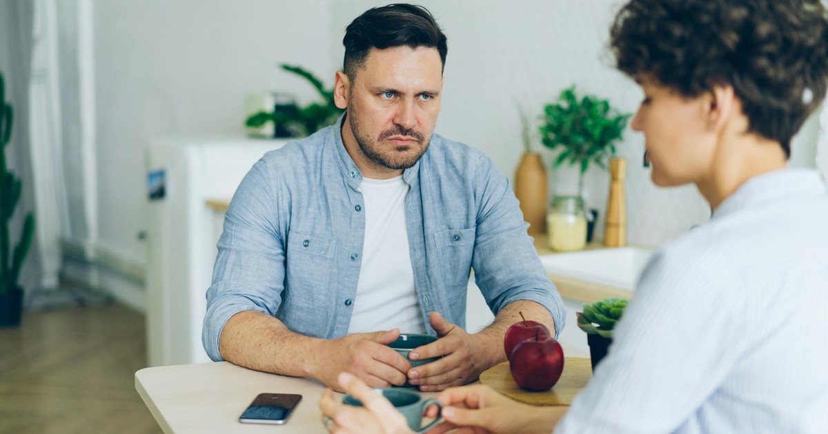 Two adults having a serious conversation at the dining table in a modern, well-lit kitchen.