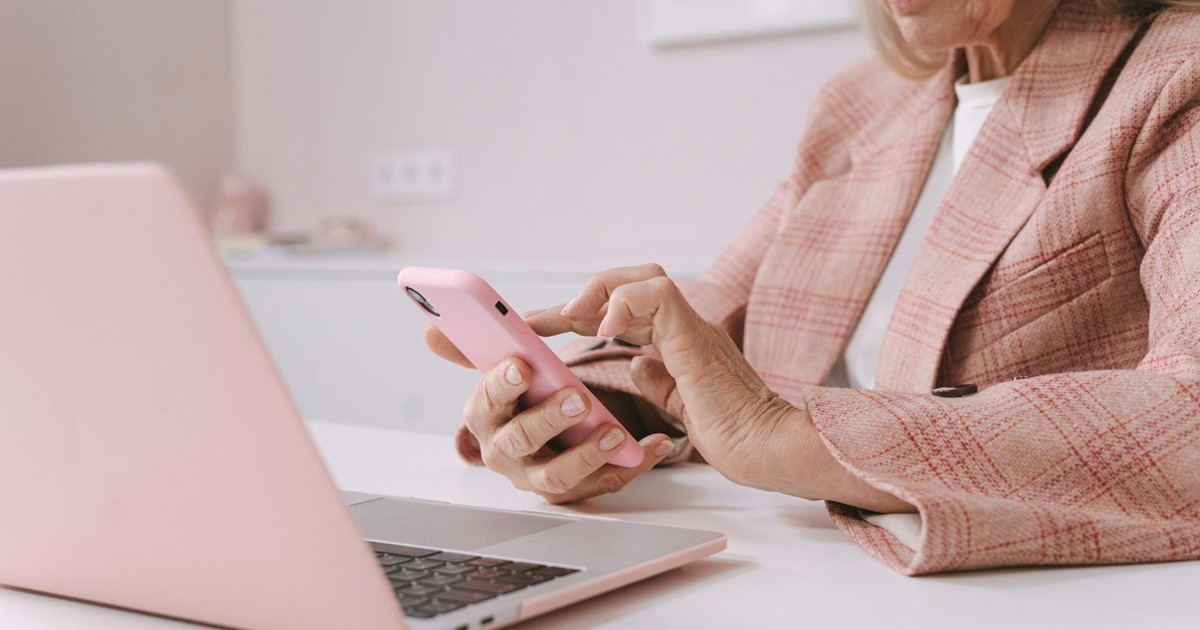 Elderly woman in pink plaid suit using a smartphone and laptop at a desk.