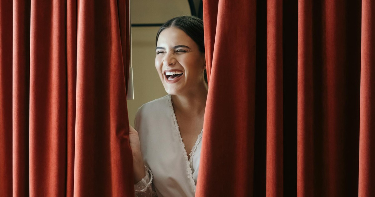 A joyful bride laughing while peeking from behind a red curtain, capturing a moment of genuine happiness.