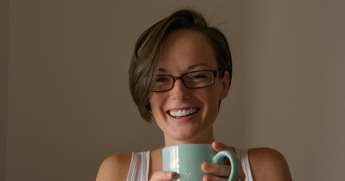 Happy woman wearing glasses and holding a mug, smiling warmly inside a cozy room.