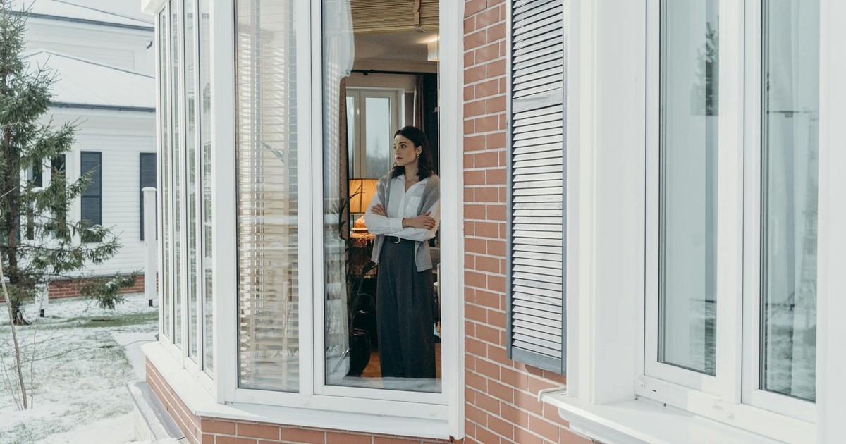 A woman stands indoors, arms crossed, looking out large windows on a snowy day, reflecting indoors and outdoors contrast.