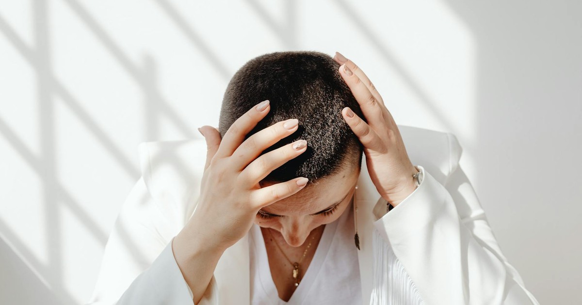 Woman in white outfit feeling stressed, sitting with hands on head in bright sunlight indoors.