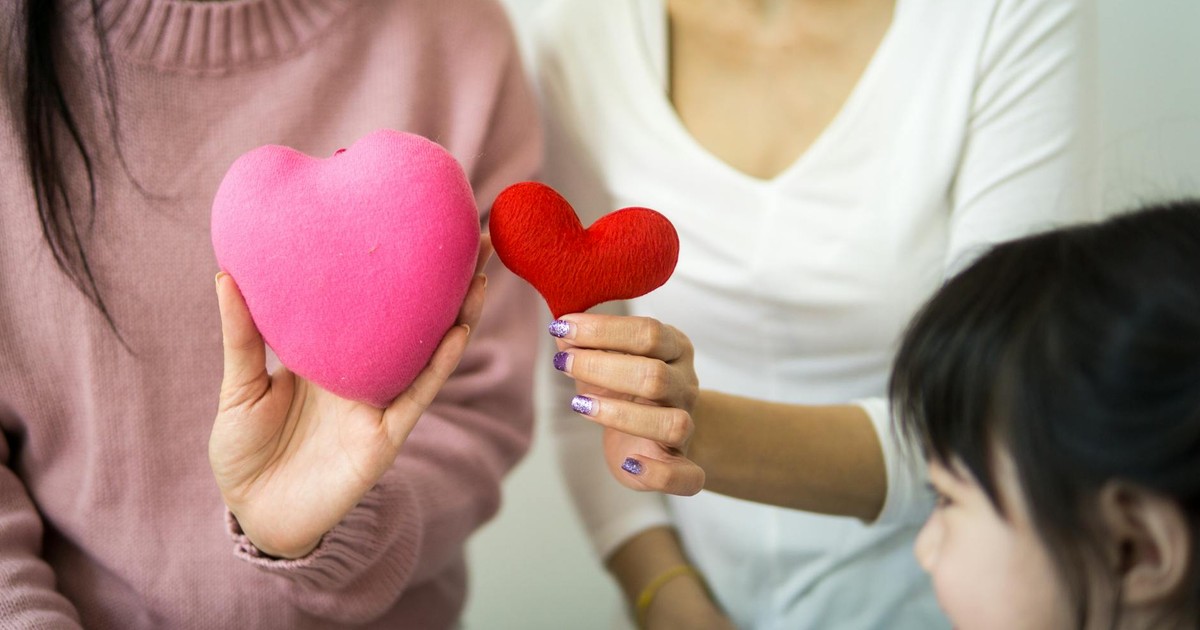 Unrecognizable ladies in casual wear demonstrating decorative hearts while sitting on couch near small kid in light room in daylight