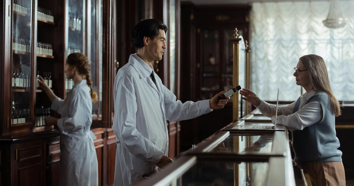 A pharmacist hands medication to a customer at a classic vintage pharmacy counter.