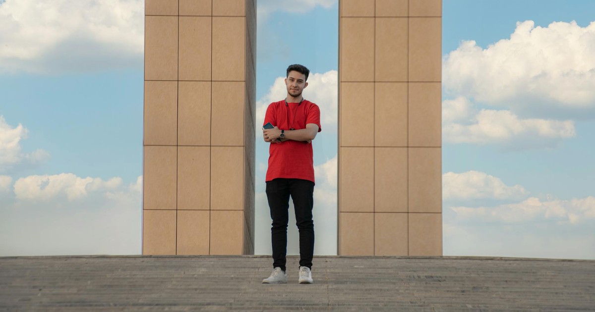 A man in a red shirt stands with arms crossed in front of a modern structure under a cloudy sky.