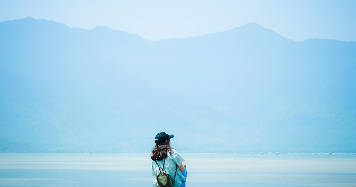 A person sitting alone by a lake, with mountains in the background, under a bright sky.