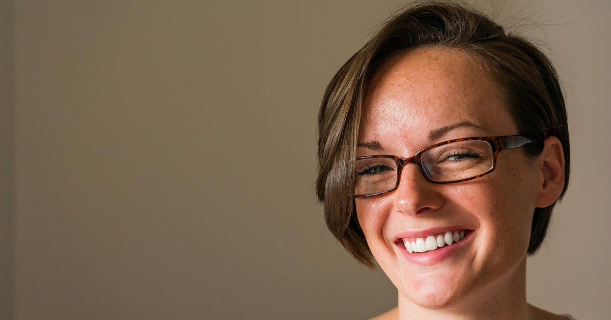 A cheerful woman with short hair and glasses smiles warmly indoors, exuding positivity.