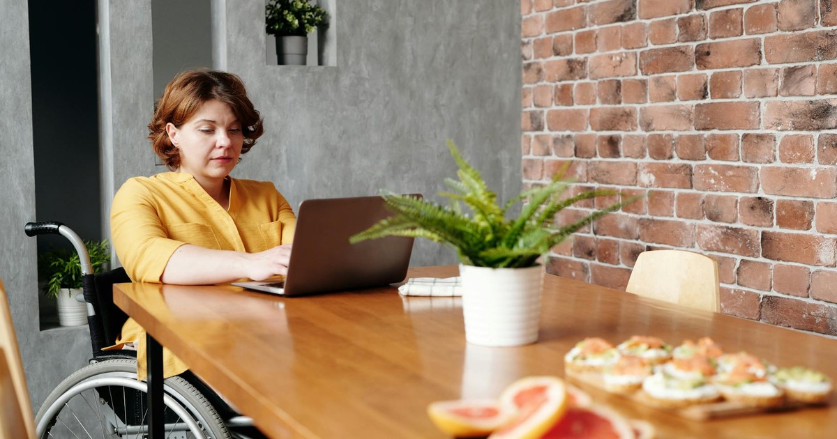 Woman working remotely on laptop in wheelchair at home dining area.