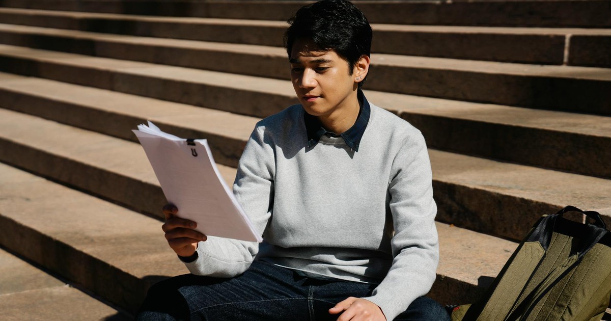 A young man sitting on stairs outdoors, studying papers in the sunlight.