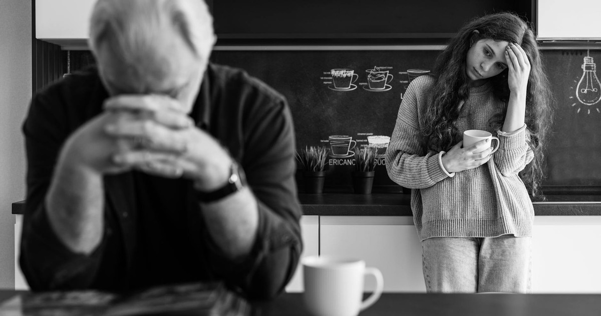A man and woman in a kitchen reflect with mugs, conveying emotions.