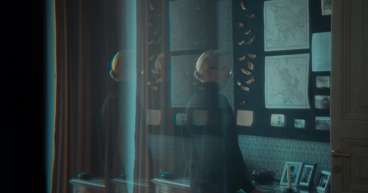 A woman with glasses stands in a dim room examining vintage maps. The scene is calm and introspective.