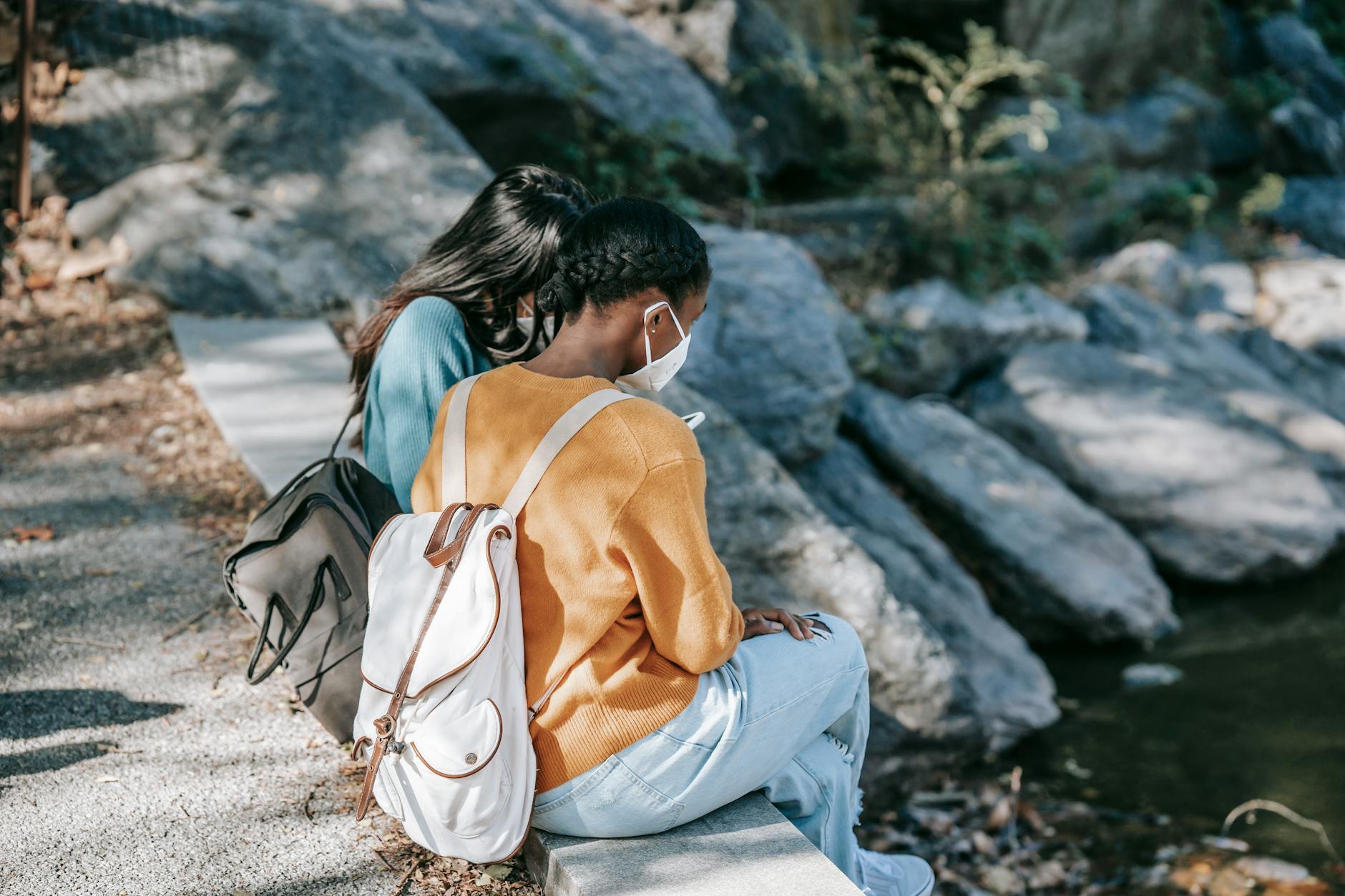 Back view of anonymous diverse friends resting on step near pond with stones in peaceful park