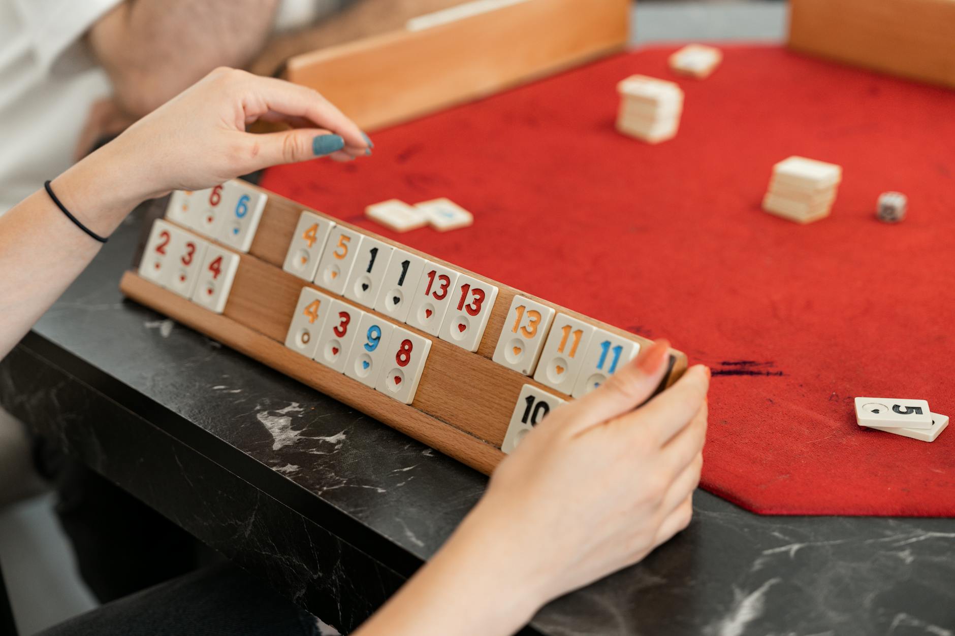 Close-up of a hand arranging Rummikub tiles on a red felt table indoors.