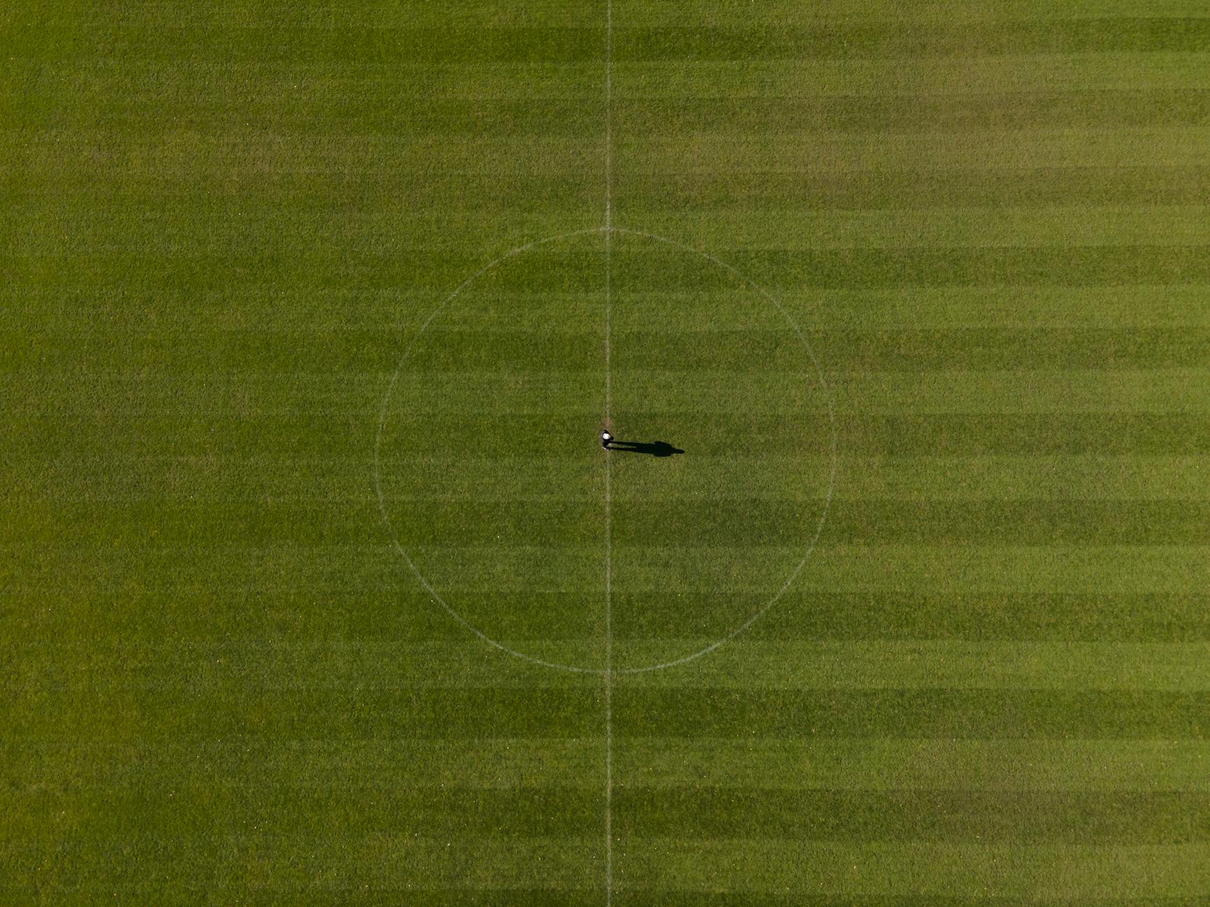 Drone shot of a soccer field with one person at the center circle, highlighting the vast green expanse.
