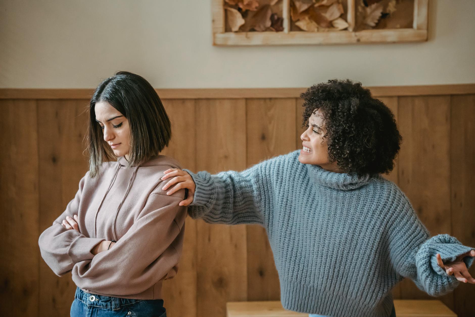 Unhappy young multiethnic female friends in casual outfit arguing with hand on shoulder while standing in bright house near wall
