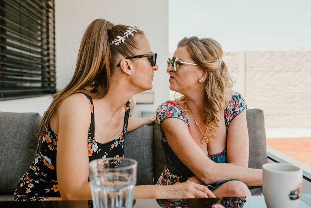 Two women playfully make faces at each other while sitting outdoors in summer