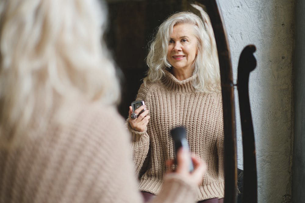 A senior woman in a cozy sweater smiles while applying makeup in front of a mirror