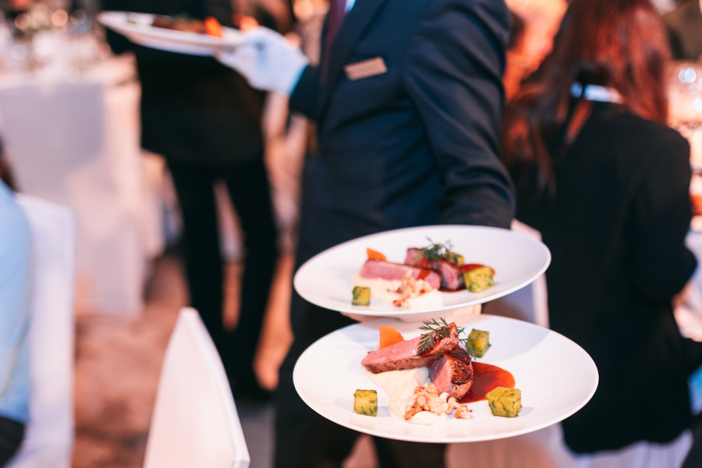 Two gourmet plates with sliced meat, vegetable cubes, sauce, and purée being served by a white-gloved waiter during a formal dinner gathering.