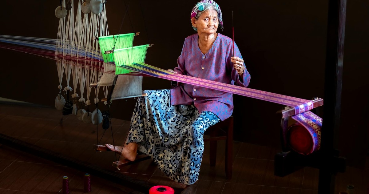 Elderly woman weaving colorful fabric on a traditional loom indoors.