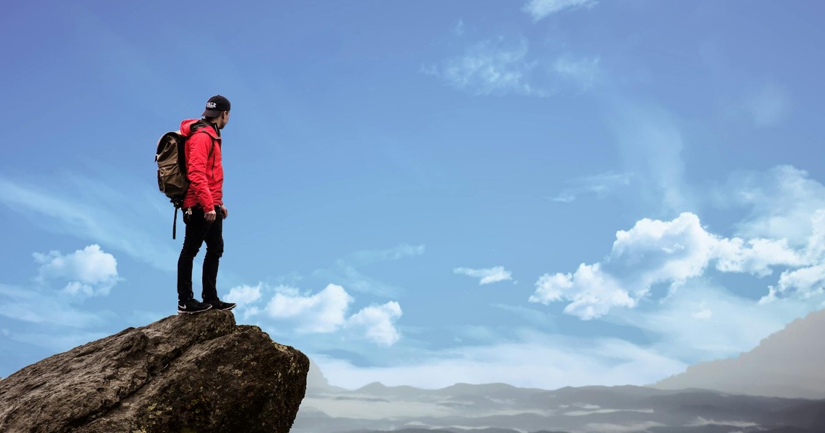 Hiker in red jacket stands on a cliff edge, overlooking a scenic mountain view under a bright blue sky.