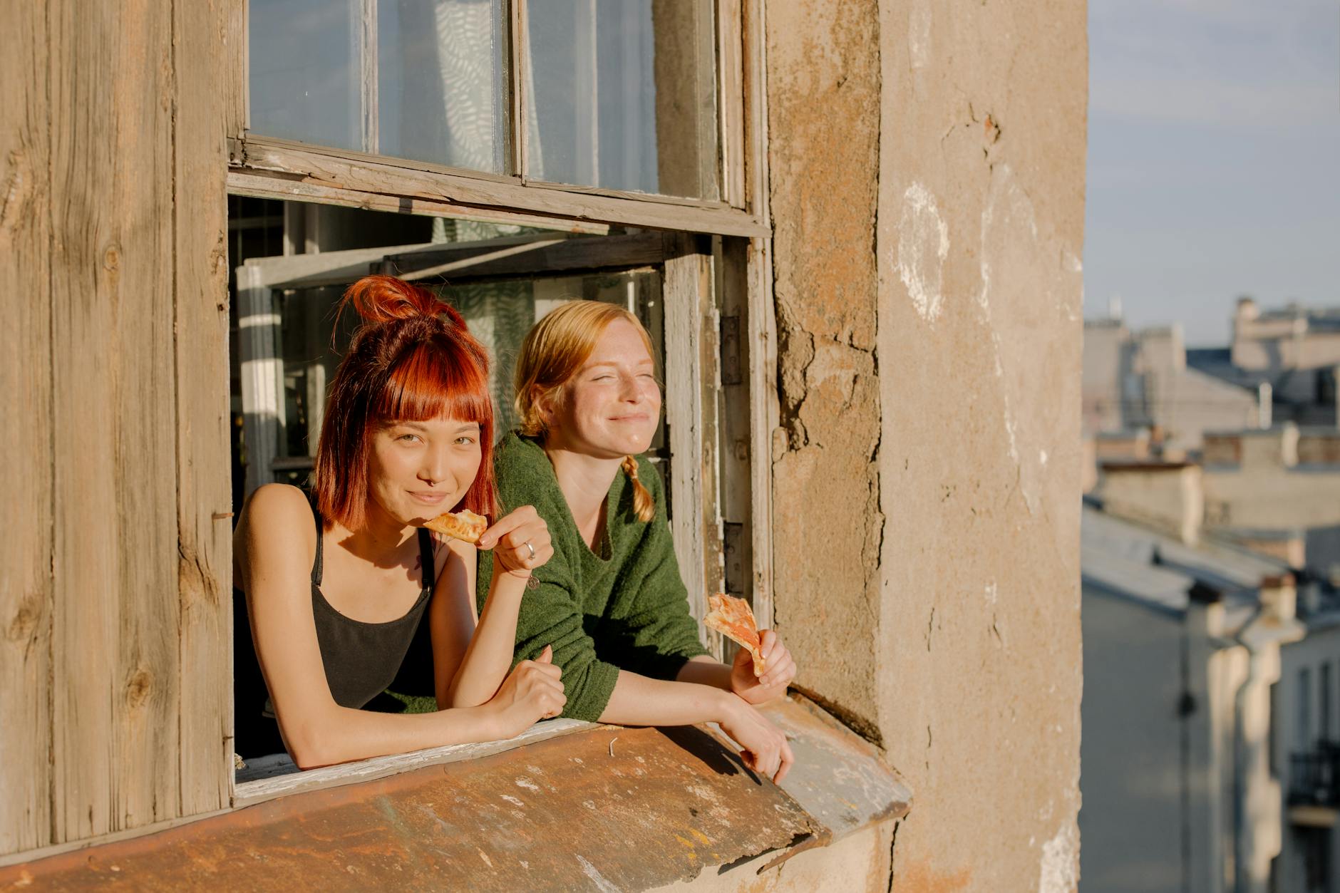 Two young women enjoying pizza by a window in a sunny urban environment.