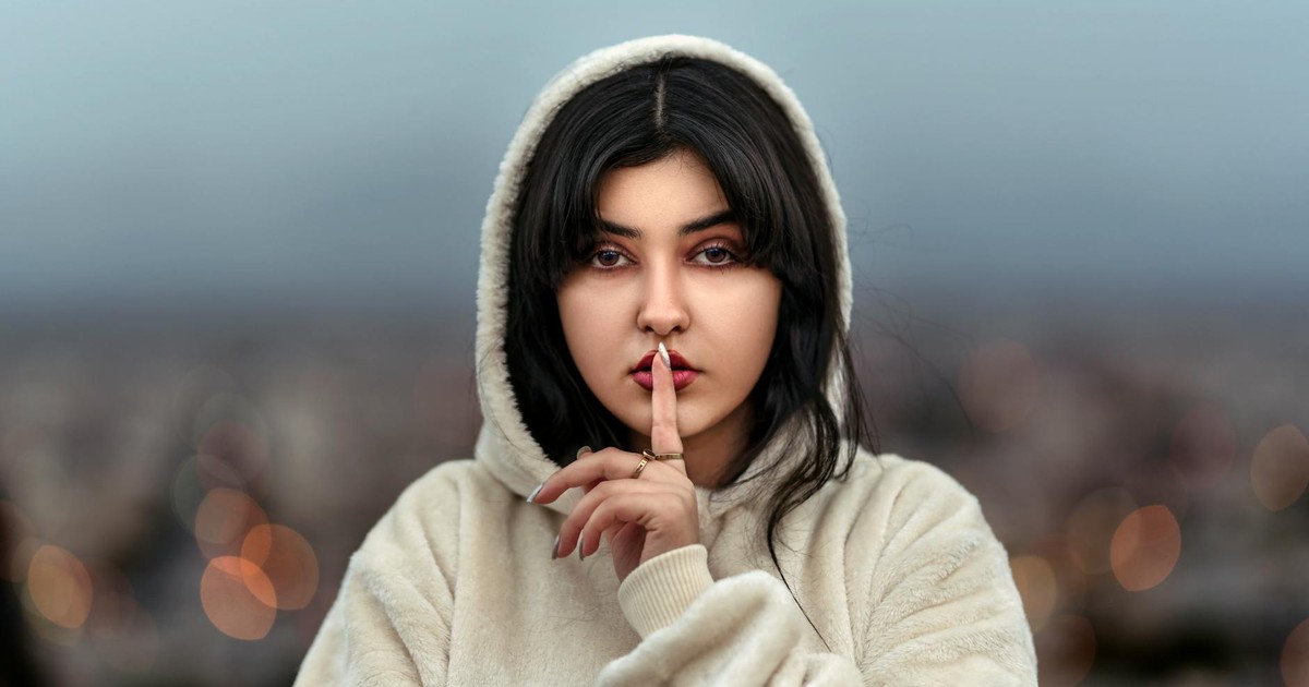 Middle Eastern woman in a hoodie signals silence, standing outdoors with an evening backdrop.