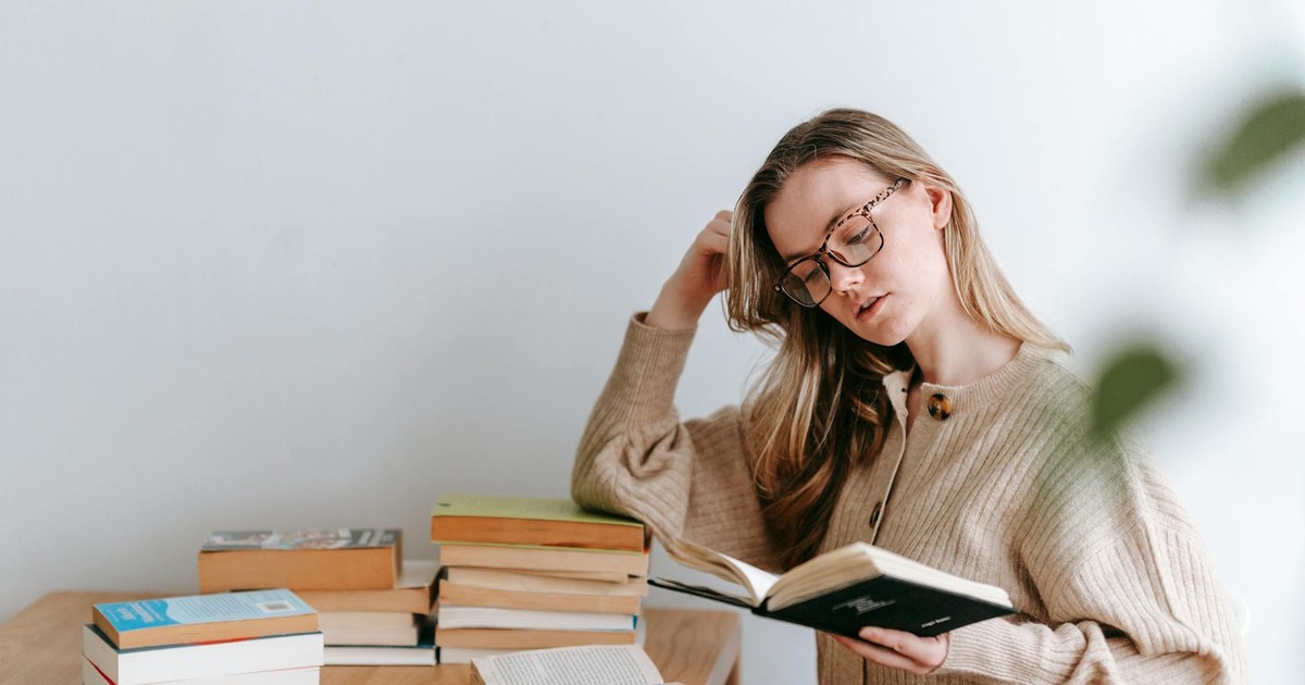 Thoughtful female student in sweater and eyeglasses standing and reading book in light room