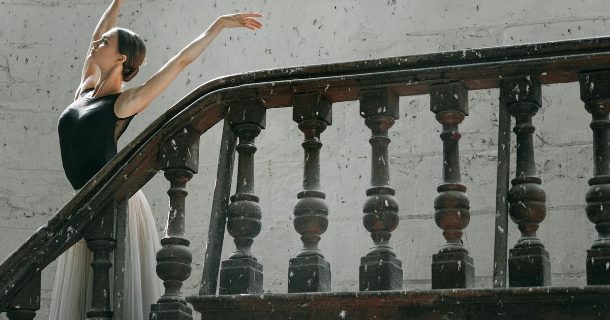A graceful ballerina poses elegantly with hands raised beside a vintage staircase.