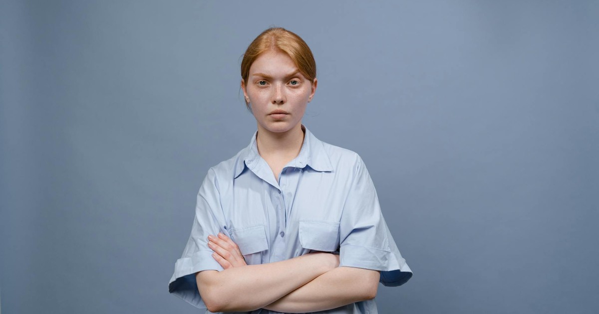 A young woman standing with crossed arms against a plain blue background, expressing annoyance.