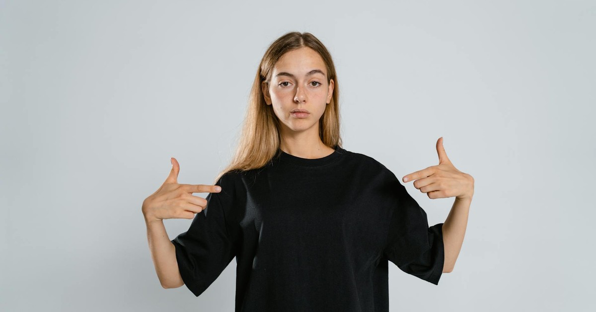 Portrait of a woman in a black t-shirt pointing at herself on a white background.