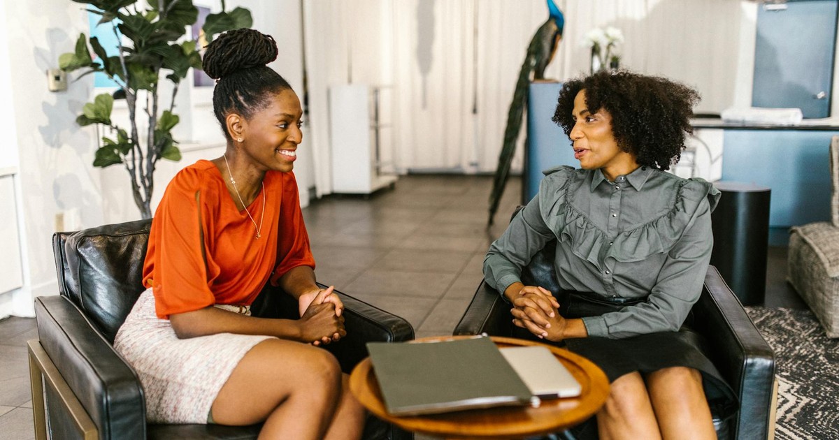 Two professional women sitting in a modern office setting, engaged in a friendly conversation.