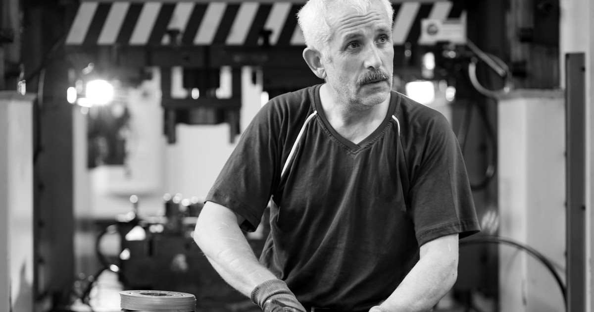 Black and white photo of an elderly worker with gloves in a workshop setting.