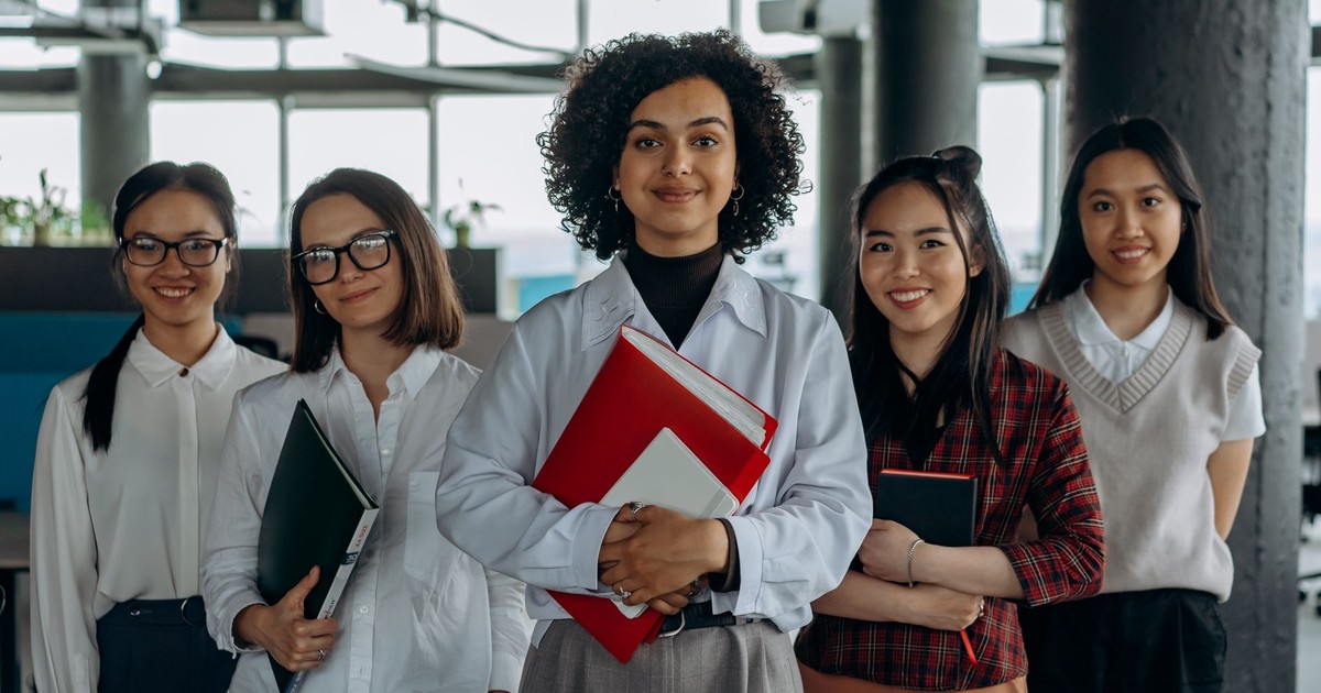 Group of diverse women colleagues smiling in a modern office setting, holding documents.