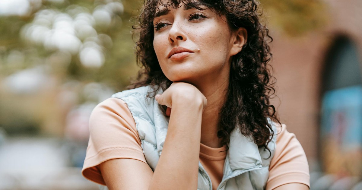 Low angle of crop pensive female with dark curly hair touching chin while looking away