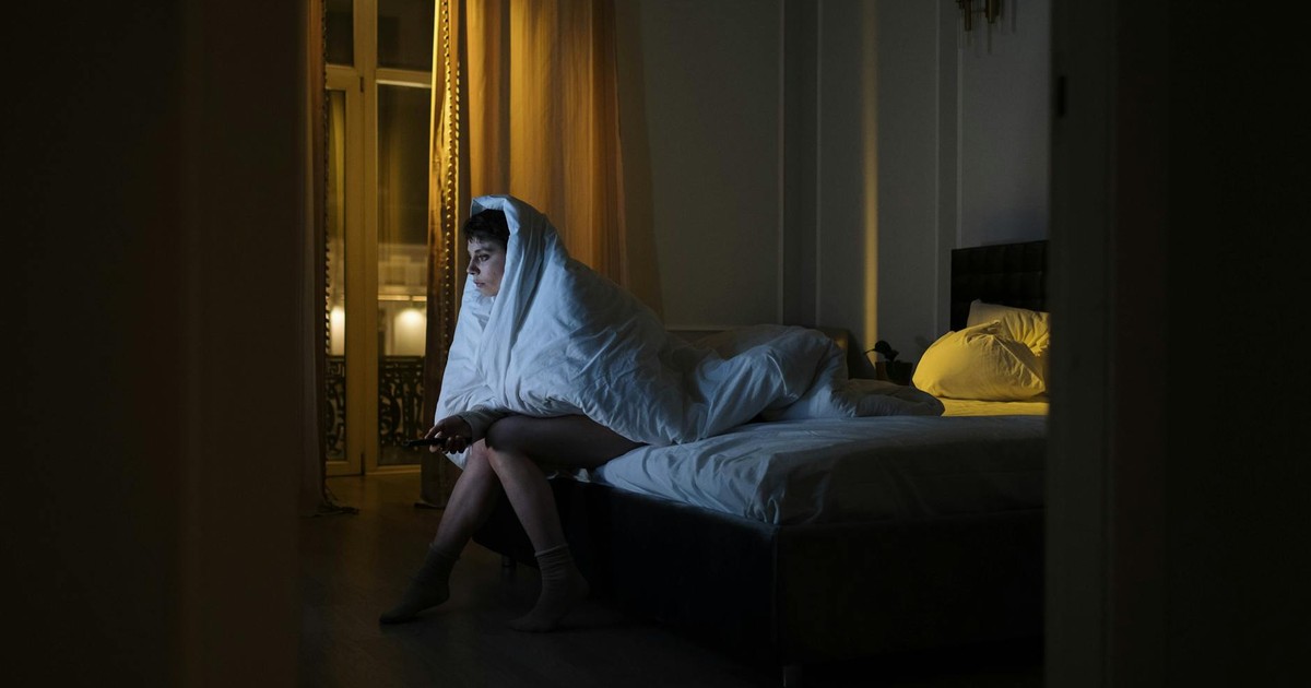 Woman wrapped in blanket watching TV, battling insomnia in dimly lit bedroom.