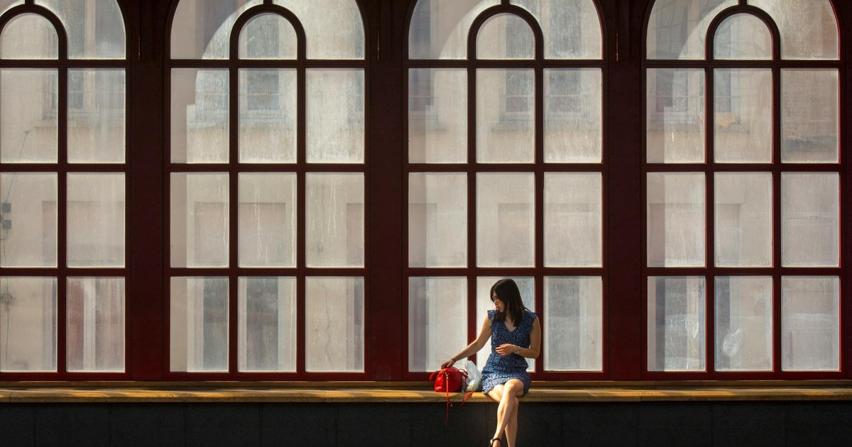 A woman sits alone in the Antwerp railway station, silhouetted by large arched windows.